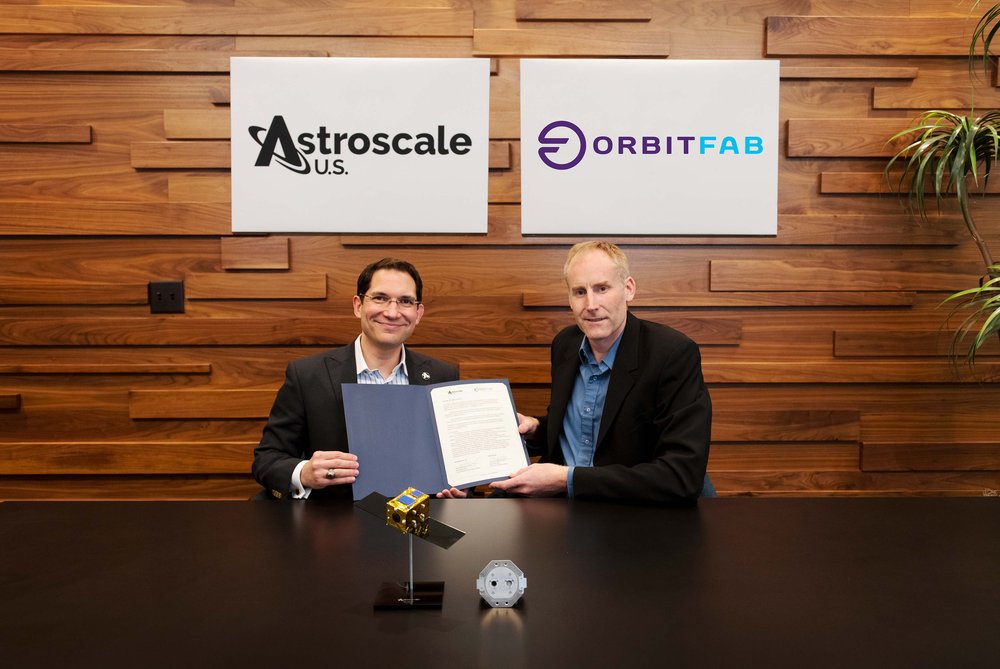 Two people holding a signed agreement with Astroscale US and Orbit Fab logos in background and Astroscale satellite and Orbit Fab's RAFTI in foreground